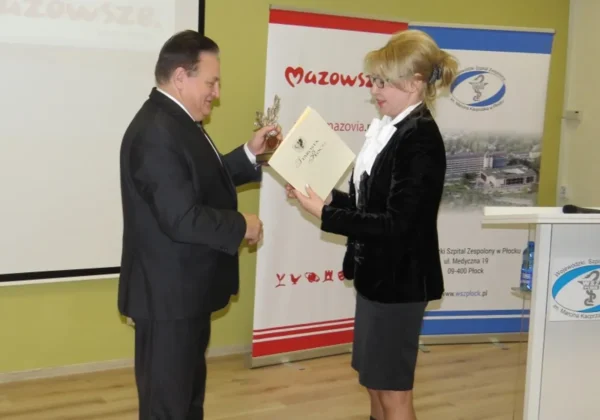 Mężczyzna w garniturze wręcza kobiecie dyplom i statuetkę podczas konferencji pt. Ekologiczne i ekonomiczne aspekty zastosowania systemu energii odnawialnej, na tle bannerów Mazowsza.