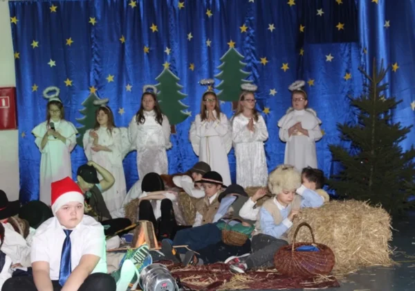 Dzieci w białych strojach aniołów stoją na scenie szopki wigilijnej w Słupnie, podczas gdy reszta uczestników w strojach pasterzy siedzi lub leży na belach siana, na tle niebieskiej sceny z gwiazdami i choinkami.