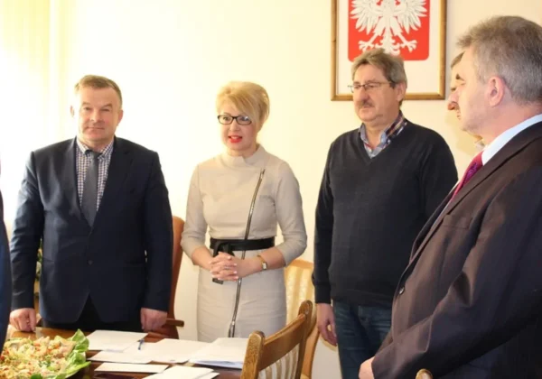 Grupa pięciu osób w formalnych ubraniach stoi przy stole z dokumentami podczas świątecznego posiedzenia Prezydium Zarządu Oddziału Powiatowego ZOSP RP w Płocku, a w tle widoczny jest godło Polski.
