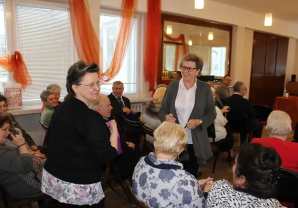 Seniorzy siedzą w sali podczas X Przeglądu Twórczości Literackiej Emerytów Rencistów i Inwalidów Regionu Płockiego, a kobieta w szarej marynarce rozmawia z nimi.