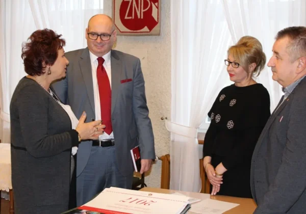 Cztery osoby w eleganckich ubraniach rozmawiają przy stole w sali z logo ZNP w tle, omawiając dokumenty dotyczące reformy oświaty.