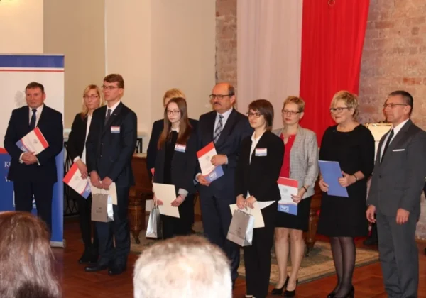 Grupa osób w formalnych strojach stoi w rzędzie podczas ceremonii wręczania stypendiów Prezesa Rady Ministrów RP, trzymając dokumenty i kolorowe teczki.