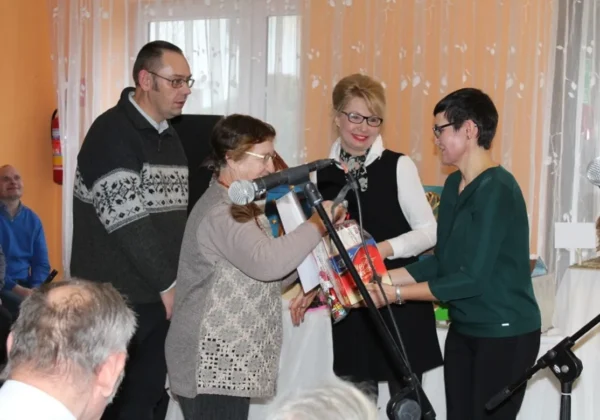 Grupa osób na scenie podczas świątecznego spotkania, kobieta w zielonej bluzce wręcza prezent innej kobiecie przy mikrofonach, w tle uśmiechnięci uczestnicy.
