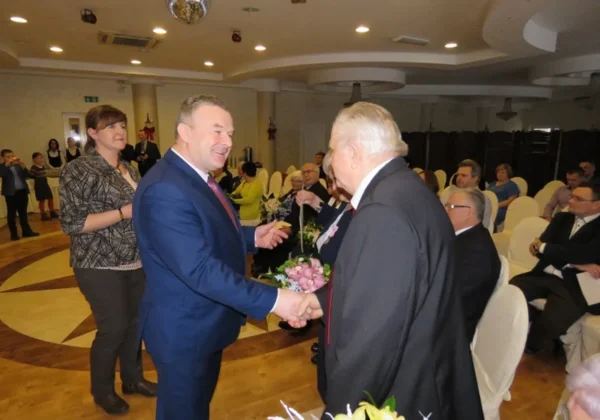 Mężczyzna w niebieskim garniturze wręcza kwiaty starszemu mężczyźnie podczas ceremonii wręczania medali małżeństwom z gminy Słupno w sali konferencyjnej.