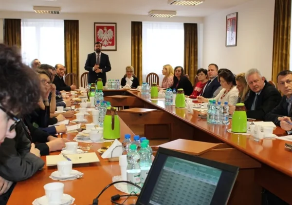 Spotkanie samorządowców w sali konferencyjnej, stojący mówca przemawia do uczestników siedzących przy długim stole; na ścianach godło Polski, a na stole znajdują się notatniki, filiżanki, butelki wody i dzbanki.