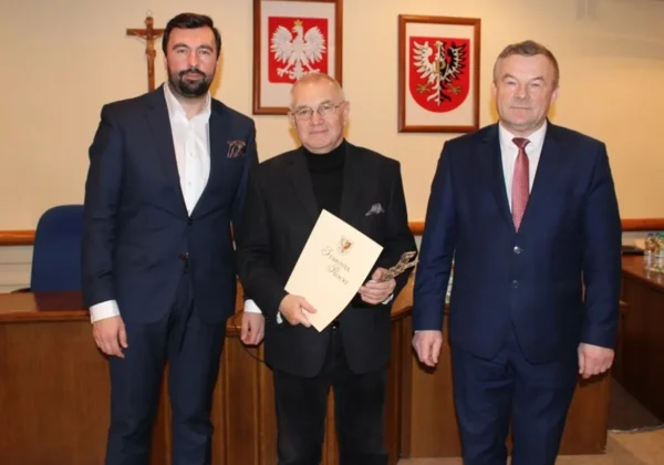 Trzech mężczyzn w garniturach stoi na sali obrad podczas XXI sesji Rady Powiatu, a w tle widoczne są herby Polski.