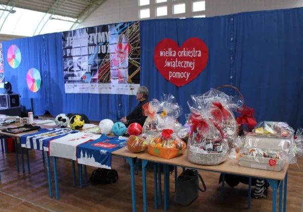 W hali Powiatu Płockiego mieszkańcy znów otwierają serca dla WOŚP; za stołem z prezentami siedzi osoba, a na stole leżą piłki i kosze upominków, w tle błękitne kurtyny i czerwone serce z napisem Wielka Orkiestra Świątecznej Pomocy.