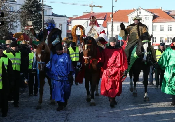 Orszak Trzech Króli przemierza ulicę miasta; królowie na koniach w kolorowych szatach prowadzeni przez uczestników, otoczony przez widzów i osoby w odblaskowych kamizelkach.