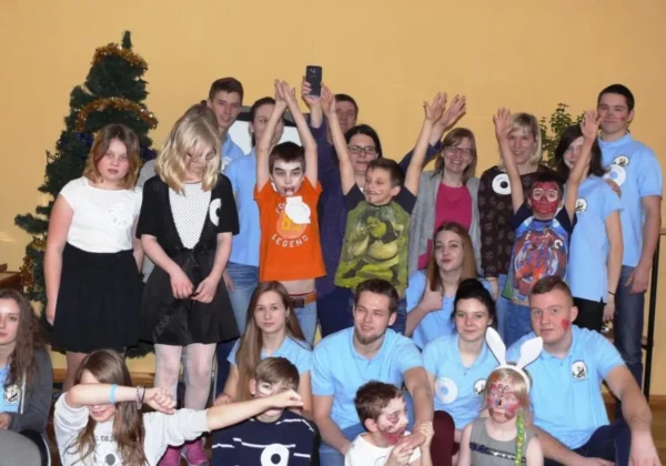 Grupa uczestników karnawałowego spotkania integracyjnego pozuje do wspólnego zdjęcia, część ubrana w niebieskie koszulki, część w przebraniach i z makijażem, na tle żółtej ściany i dekorowanej choinki.