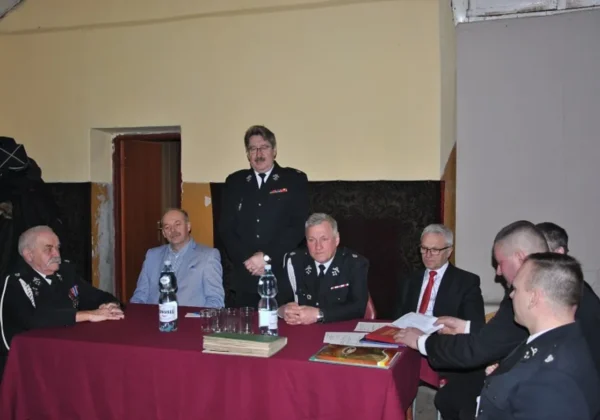 Grupa mężczyzn w mundurach strażackich i garniturach siedzi przy stole z czerwonym obrusem, omawiając dokumenty podczas półmetka kampanii sprawozdawczej Ochotniczych Straży Pożarnych.
