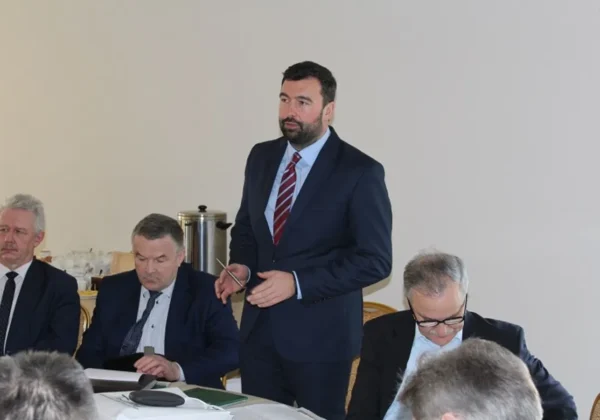 Mężczyzna w granatowym garniturze stoi i przemawia podczas Konwentu Samorządów Powiatu Płockiego, który wrócił po długiej przerwie, podczas gdy pozostali uczestnicy siedzą przy stole i przeglądają dokumenty.