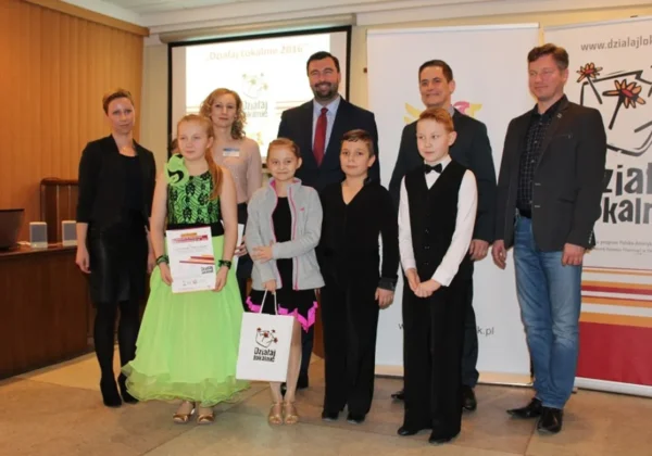 Grupa uczestników i organizatorów stoi na scenie podczas gali podsumowującej program Działaj Lokalnie IX, trzymając certyfikaty i torbę z logo programu.