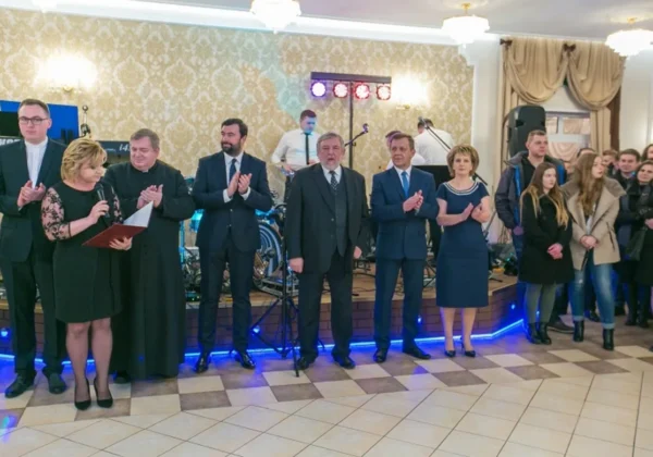Grupa osób w eleganckich strojach stoi w sali balowej podczas studniówki, kobieta trzyma mikrofon i kartkę, a w tle gra zespół muzyczny.