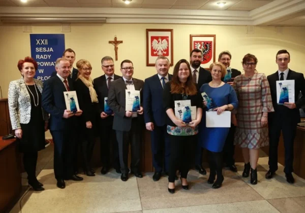Grupa osób w eleganckich strojach stoi w sali sesyjnej Rady Powiatu podczas XXII sesji, trzymając nagrody i dyplomy jako wyróżnienia dla wyjątkowych osób.