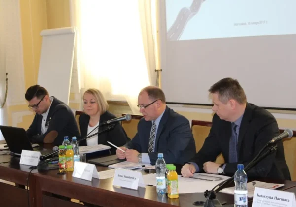 Czterech uczestników spotkania z beneficjentami w garniturach siedzi przy długim stole konferencyjnym, omawiając materiały dotyczące dróg lokalnych w 2017 roku, przy mikrofonach i napojach na stole.