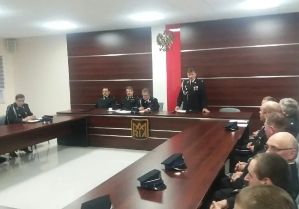Spotkanie strażaków w mundurach w sali konferencyjnej podczas kampanii sprawozdawczej OSP; prelegent stoi przy mównicy, a pozostali uczestnicy siedzą przy stołach, z widocznym polskim godłem i biało‑czerwonym motywem w tle.