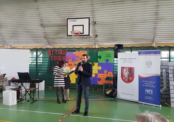 Na hali sportowej podczas XI Targ Edukacji i Pracy w Małej Wsi mężczyzna przemawia do mikrofonu na scenie, obok niego stoją dwie kobiety, a w tle widać kolorowe banery i stanowiska wystawowe.