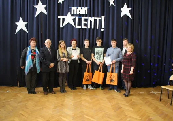 Grupa osób na scenie podczas wydarzenia Mam Talent w Wyszogrodzie, przed czarną kurtyną z gwiazdami i napisem Mam Talent, trzymająca certyfikaty i pomarańczowe torby.