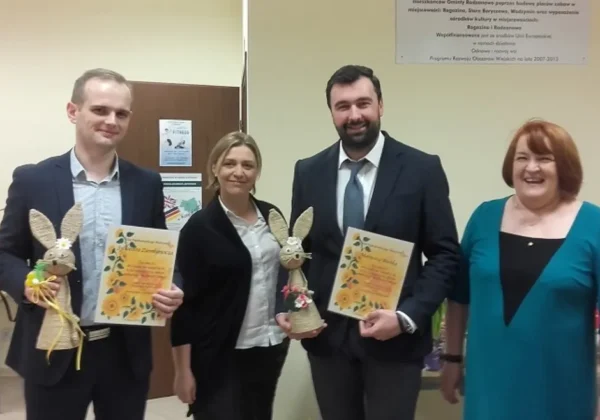 Cztery osoby stoją w biurowym holu: dwóch mężczyzn w garniturach i dwie kobiety, wszyscy uśmiechnięci, trzymają certyfikaty oraz ozdobne zajączki.