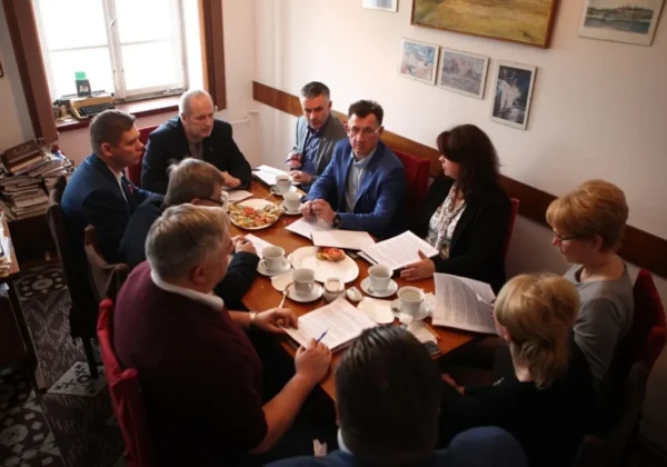 Grupa osób przy stole w biurze omawia dokumenty na spotkaniu dotyczącym inwestycji z Tumskiego Wzgórza; na stole stoją filiżanki i przekąski.