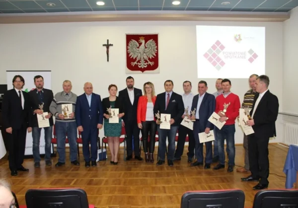 Grupa liderów stojących na sali konferencyjnej w Drobinie, trzymających statuetki i certyfikaty podczas spotkania, z krzyżem, herbem powiatu i projekcją z napisem „Powiatowe Spotkanie” w tle.
