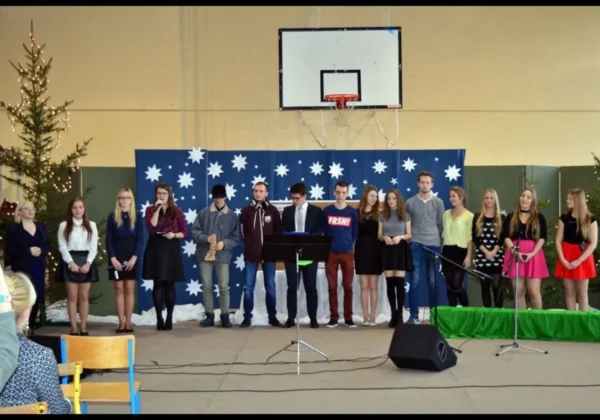 Grupa uczniów Zespołu Szkół w Gąbinie stoi na scenie w szkolnej hali podczas występu, przed niebieską dekoracją z białymi gwiazdami i choinkami.
