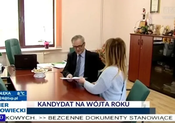 Dwóch dorosłych w biurze – mężczyzna w garniturze podpisuje dokumenty, kobieta podaje mu papiery, a w tle wyświetla się informacja o głosowaniu na Wójta Roku.