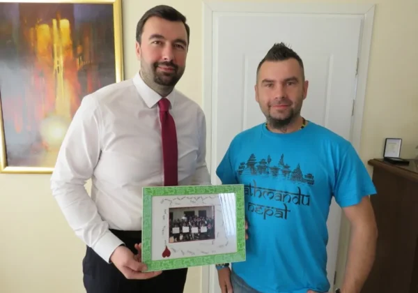 Dwóch mężczyzn stoi w pomieszczeniu, jeden w białej koszuli i czerwonym krawacie, drugi w turkusowej koszulce, a razem trzymają zieloną ramkę z fotografią grupową.
