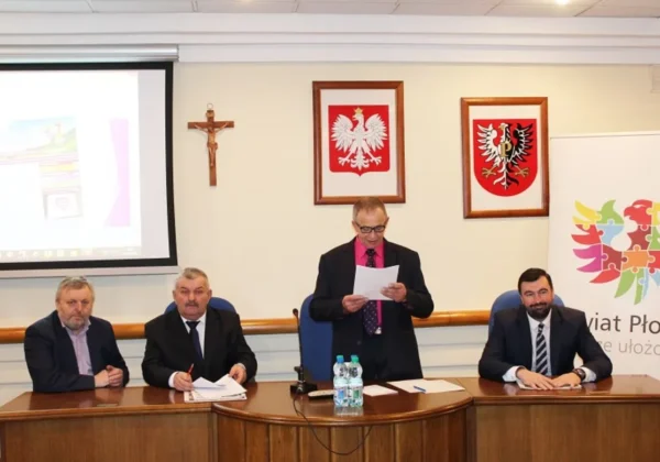Czterech mężczyzn siedzi przy stole konferencyjnym podczas wspólnego posiedzenia rolników powiatów płockiego i sierpeckiego, z prezentacją na projektorze po lewej i dwoma herbami na ścianie w tle.
