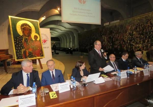 Panel dyskusyjny podczas XXI Nadzwyczajnego Zgromadzenia Ogólnego Związku Powiatów Polskich: mężczyzna przemawia z mikrofonem przy stole, w tle obraz Matki Bożej z Dzieciątkiem i projekt na ekranie.