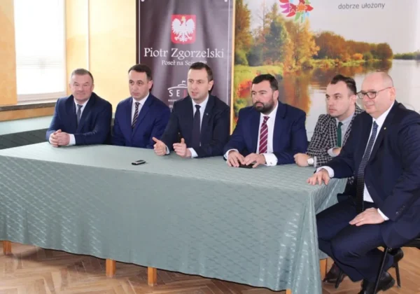 Grupa sześciu mężczyzn w garniturach siedzi za stołem podczas konferencji prasowej w Powiecie Płockim, promującej politykę senioralną, z banerami w tle.