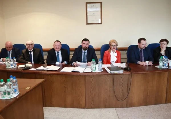 Grupa siedmiu urzędników i radnych w formalnych ubraniach siedzi przy długim drewnianym stole w sali sesyjnej podczas Sesji Rady Powiatu, z dokumentami i butelkami wody przed sobą.