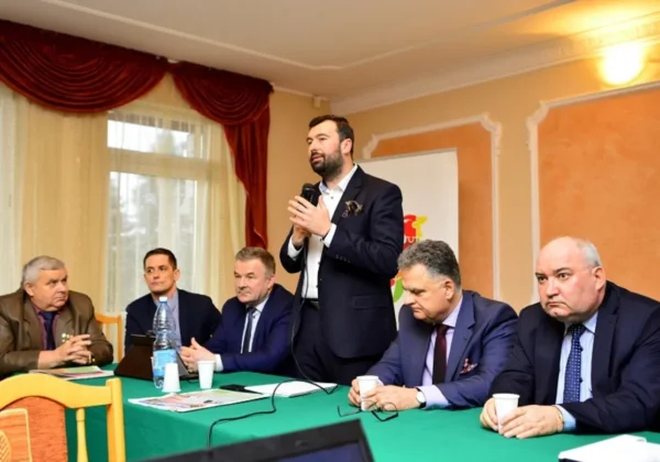 Grupa mężczyzn w garniturach siedzi przy zielonym stole podczas powiatowego spotkania liderów lokalnych; jeden z nich stoi z mikrofonem i przemawia do uczestników.