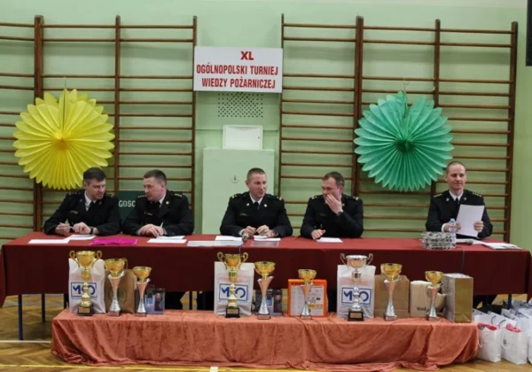 Grupa strażaków w mundurach siedzi za czerwonym stołem z nagrodami i pucharami podczas ceremonii eliminacji powiatowych turnieju pożarniczego, a w tle wisi baner XL Ogólnopolski Turniej Wiedzy Pożarniczej.