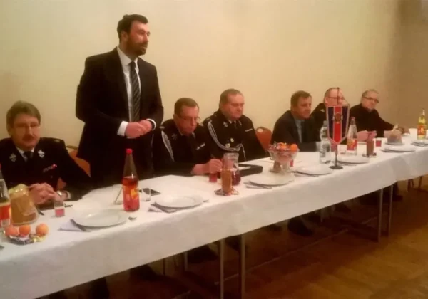 Grupa strażaków OSP w mundurach siedzi przy długim stole zastawionym napojami i jedzeniem, a stojący mężczyzna w garniturze wygłasza przemówienie.