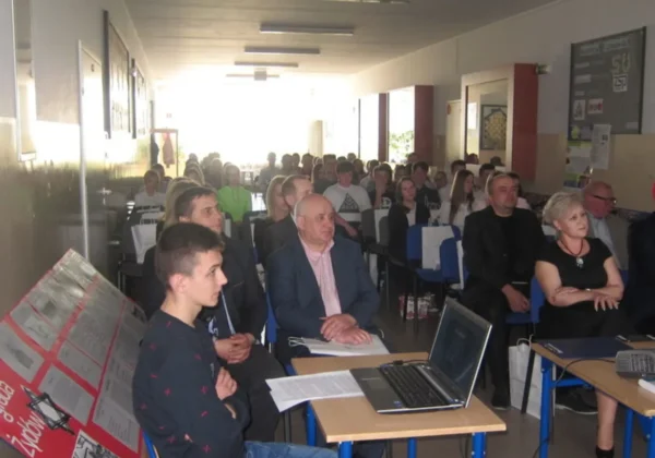 Uczestnicy spotkania w szkolnej auli Zespołu Szkół Ponadgimnazjalnych w Drobinie siedzą w rzędach na niebieskich krzesłach, a na pierwszym planie widoczny jest laptop i dokumenty na stole.