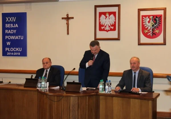 Trzech mężczyzn w garniturach siedzi za biurkiem podczas XXIV sesji Rady Powiatu w Płocku; jeden z nich stoi i przemawia do mikrofonu, w tle widoczne krzyż oraz dwa herby, a z lewej strony banner z napisem XXIV Sesja Rady Powiatu w Płocku 2014-2018.