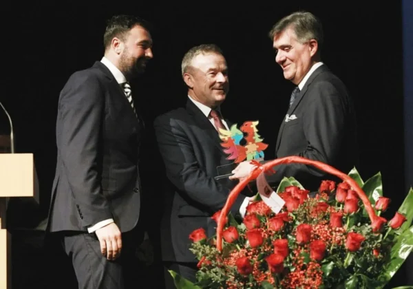 Na scenie trzech mężczyzn w garniturach podczas jubileuszu Politechniki; jeden wręcza drugiemu kolorowy puchar, a obok stoi duży bukiet czerwonych róż.