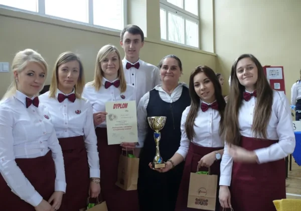 Grupa uczniów Zespołu Szkół im. Stanisława Staszica w Gąbinie, ubranych w białe koszule i burgundowe muszki, wraz z opiekunką stoi w sali i trzyma puchar oraz dyplom po zdobyciu III miejsca w konkursie „Stół ludowy we współczesnej restauracji”.