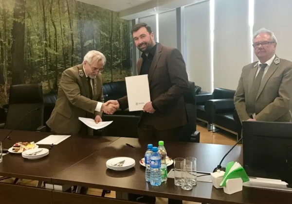 Trzech mężczyzn w garniturach w sali konferencyjnej Dyrekcji Generalnej Lasów Państwowych; dwóch z nich ściska sobie dłonie nad podpisaniem umowy przyrzeczenia, trzeci stoi z boku; na stole są napoje i przekąski, w tle widać las.