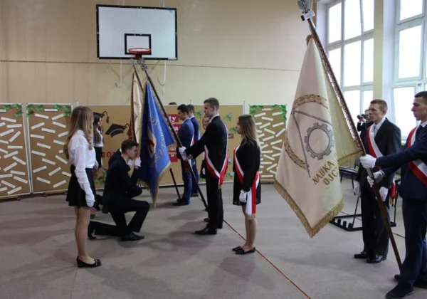 Uczniowie w eleganckich ubraniach stoją w sali gimnastycznej podczas ceremonii rozdania świadectw absolwentom ZS im. St. Staszica w Gąbinie, trzymając flagi.