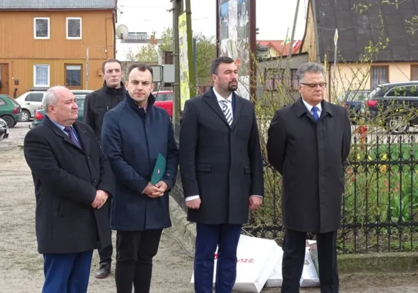 Grupa mężczyzn w ciemnych płaszczach stoi na zewnątrz przy ogrodzeniu w Bodzanowie podczas obchodów 110-lecia działalności OSP.