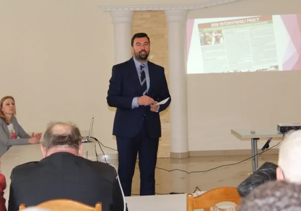 Mężczyzna w garniturze przemawia do zgromadzonych podczas spotkania z liderami gminy Słubice, po lewej stronie siedzi kobieta przy stole, a na ścianie wyświetlana jest prezentacja.