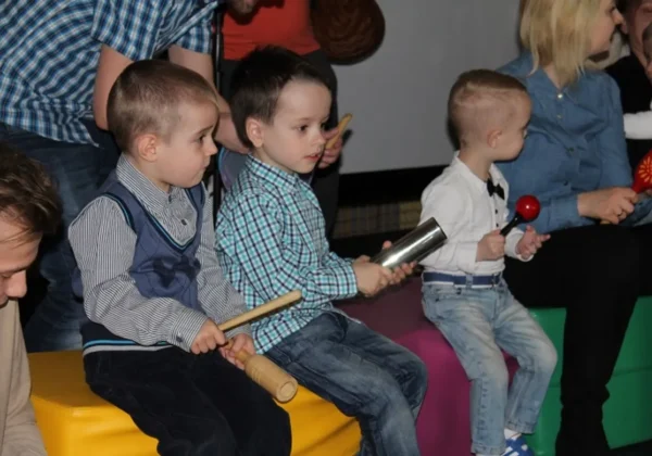 Dzieci siedzą na kolorowych pufach i grają na instrumentach perkusyjnych podczas zajęć wspomagających rozwój dziecka.