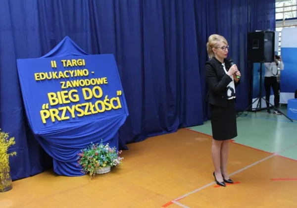 Kobieta w czarnym stroju z mikrofonem przemawia na scenie podczas II Targów Edukacyjno‑Zawodowych „Bieg do przyszłości”, obok niebieskiej dekoracji z żółtym napisem i kwiatami.