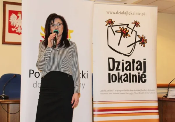 Kobieta w pasiastej bluzce trzyma mikrofon i przemawia na scenie, stojąc obok banerów programu Działaj Lokalnie.
