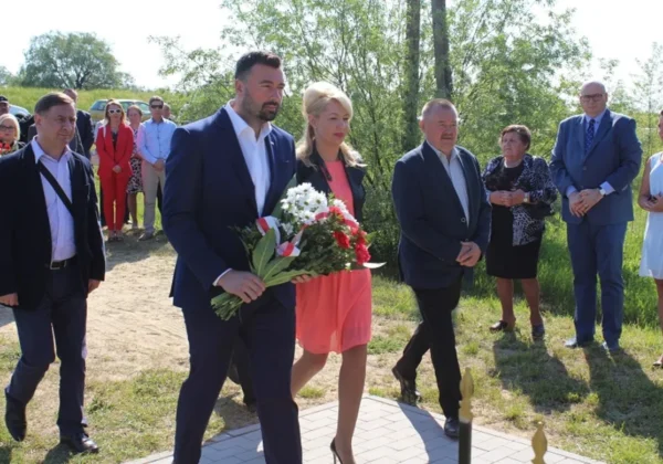 Grupa osób w formalnych ubraniach stoi na zewnątrz podczas obchodów siódmej rocznicy powodzi; mężczyzna niesie bukiet kwiatów, a reszta wygląda na zaniepokojoną obawami o brak pieniędzy na pogłębianie.