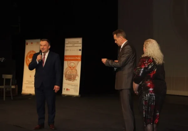 Na scenie trzech ludzi: mężczyzna w garniturze przemawia do mikrofonu, obok niego drugi mężczyzna wręcza coś kobiecie, a w tle stoją banery upamiętniające 10-lecie Banku Żywności w Płocku.