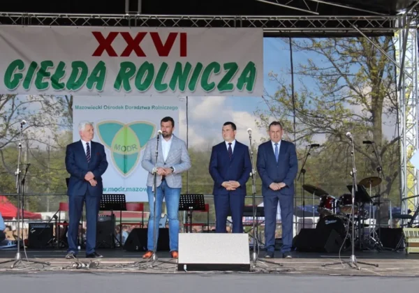 Czterech mężczyzn na scenie podczas XXVI Giełdy Rolniczej w Łącku; jeden przemawia do mikrofonu, za nimi duży baner z napisem XXVI GIEŁDA ROLNICZA.