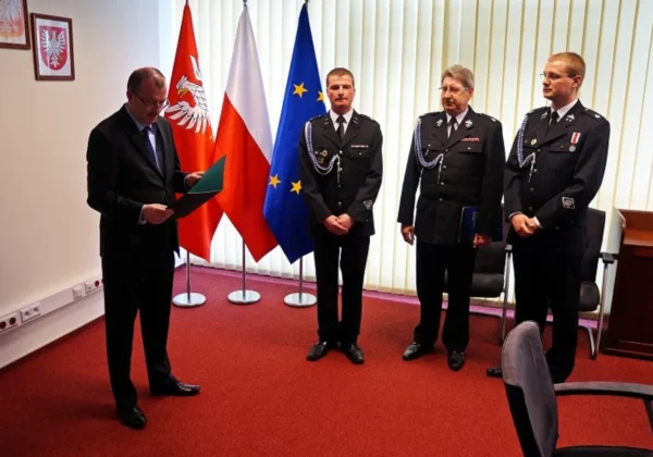 Czterech mężczyzn stoi w sali z flagami Polski, Unii Europejskiej i regionu Mazowsze; jeden w garniturze odczytuje dokument, trzech strażaków w mundurach stoi obok.